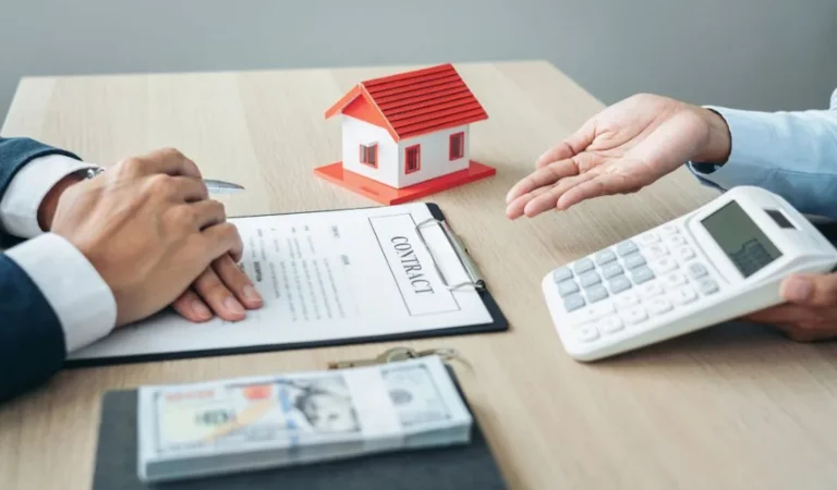 Two individuals discussing a contract with a house model, calculator, and money on a wooden table.