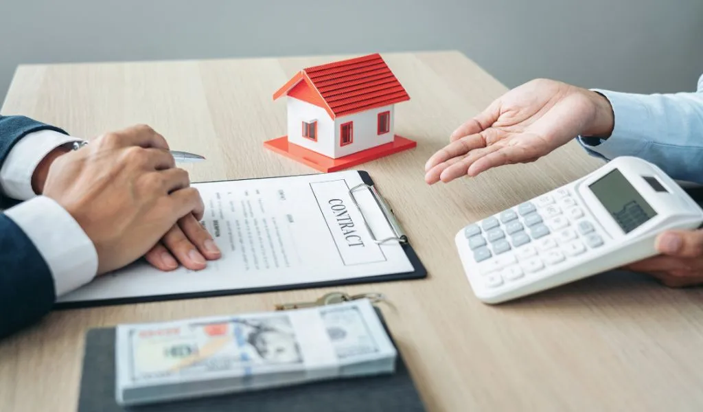 Two individuals discussing a contract with a house model, calculator, and money on a wooden table.