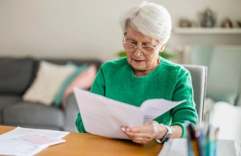 Elderly woman with white hair and glasses reading documents at wooden table indoors.