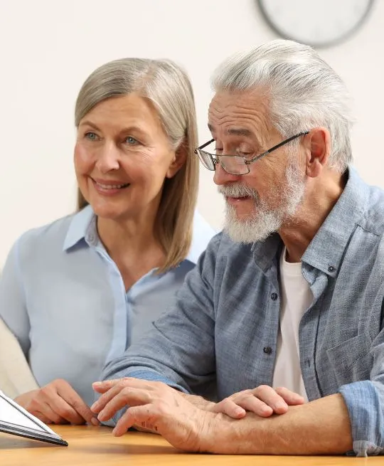 Elderly man and woman seated at table, engaging with a tablet device.