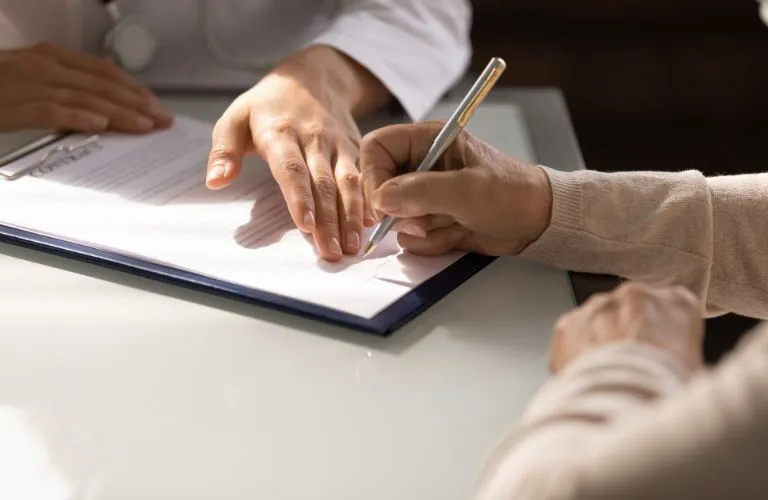 Two individuals at a desk, one signing a document with a pen, the other resting hand nearby.