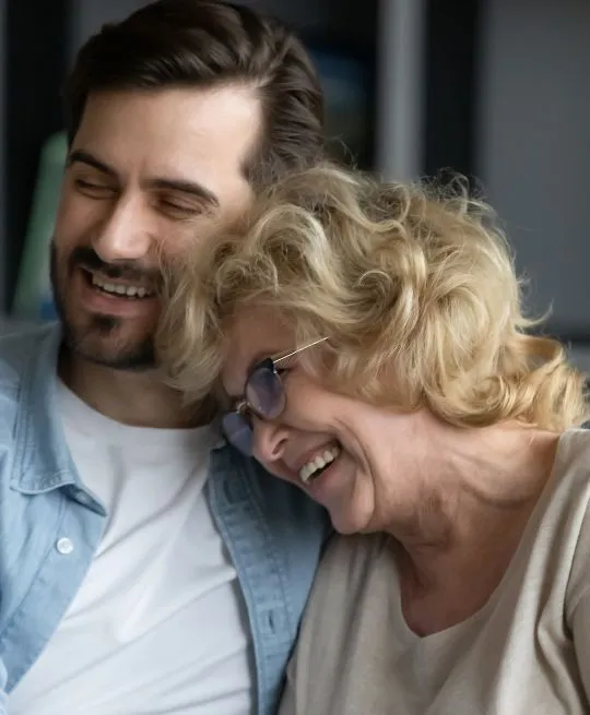 Middle-aged woman with blonde hair and eyeglasses laughing with a younger man in casual clothing.
