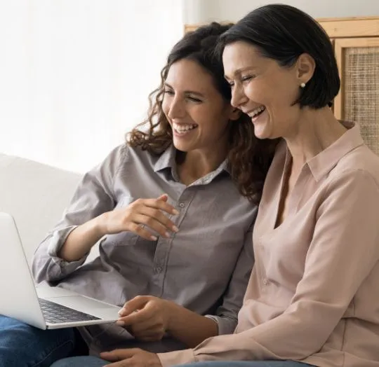 Two women sitting closely on a couch, smiling and looking at a laptop screen.