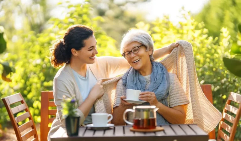Two women, one younger and one older, share a warm moment outdoors with tea, sunlight, and greenery.