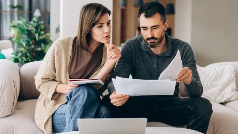 A man and woman sitting on a beige sofa reviewing documents and using a laptop.