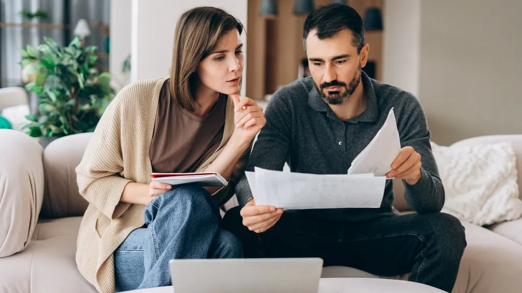 A man and woman sitting on a beige sofa reviewing documents and using a laptop.