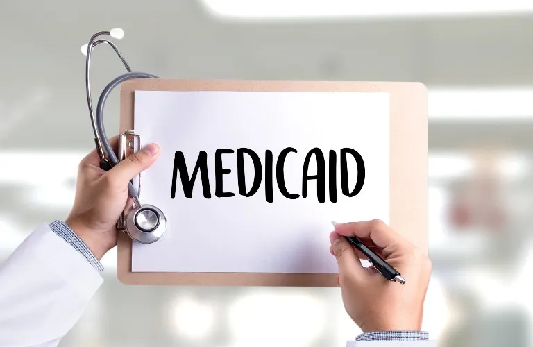 Hands holding a clipboard with the word "MEDICAID" and a stethoscope draped over it.