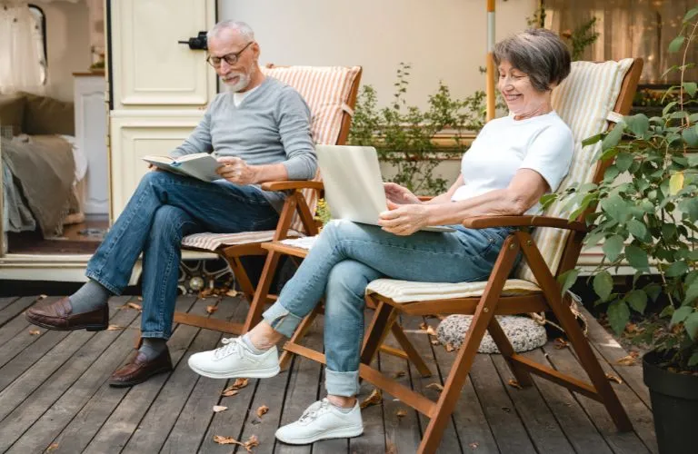 Elderly man reading a book and elderly woman using laptop, seated on wooden deck chairs outdoors.