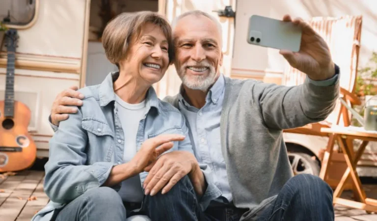 Elderly couple smiling and taking a selfie outdoors near a camper and guitar.