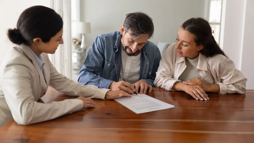 Three people seated at a wooden table reviewing and signing a document.