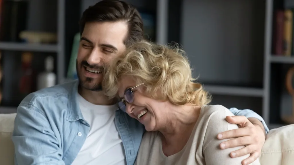 Middle-aged man and elderly woman smiling and embracing on a couch indoors.
