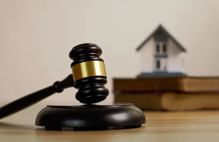Wooden judge's gavel with gold band resting on sound block, small house model and books blurred.
