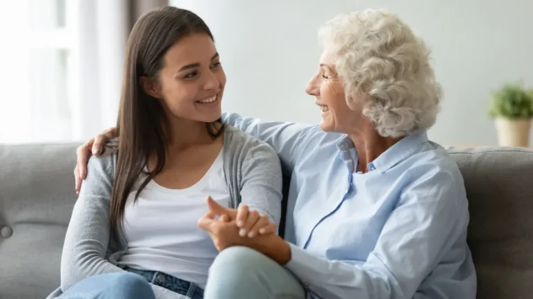 Two women, one elderly with white curly hair and one young with straight dark hair, sitting on a gray couch smiling and holding hands.