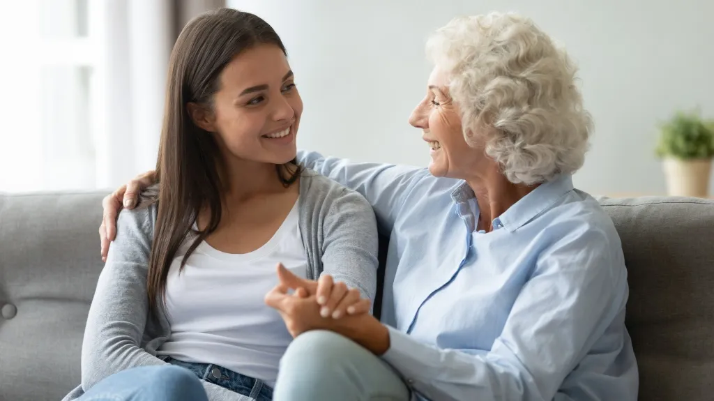 Two women, one elderly with white curly hair and one young with straight dark hair, sitting on a gray couch smiling and holding hands.