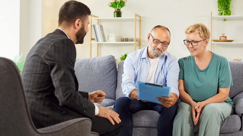 Three adults having a discussion in a modern living room, one holding a blue clipboard.