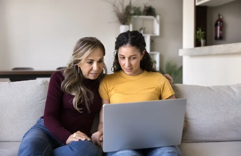 Two women sitting on a beige sofa, focused on a laptop screen together.