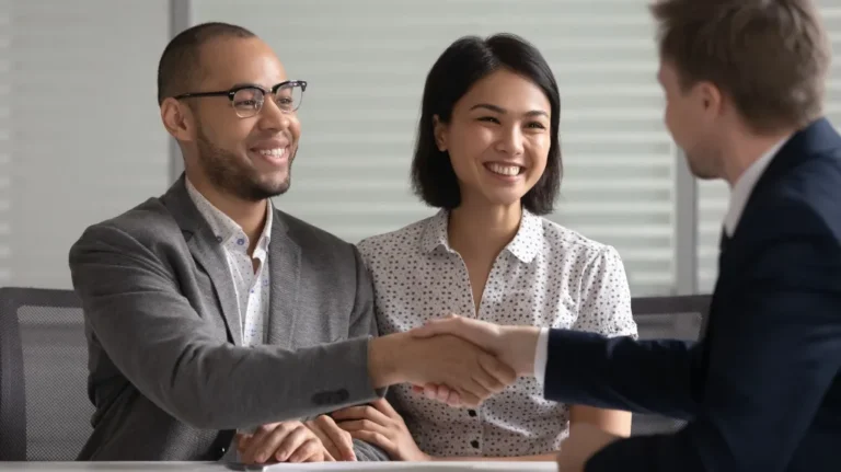 Three professionals in business attire shaking hands across a table, smiling.