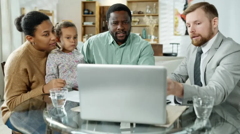 Professional man in gray suit explaining information on laptop to attentive family of three at glass table.