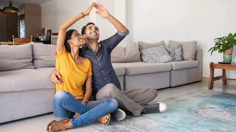 Young couple sitting on floor in living room, forming a house shape with their hands.
