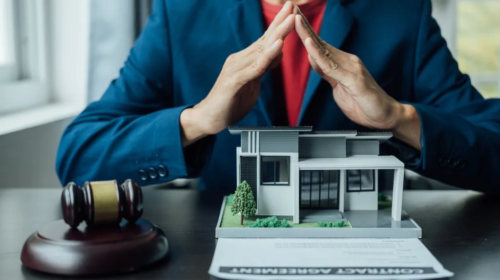 Person in blue blazer with hands forming a protective gesture over a house model and contract agreement.