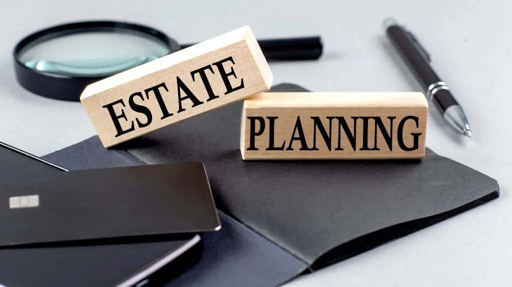 Wooden blocks with "Estate" and "Planning" text, magnifying glass, pen, credit card, and black folder on white surface.