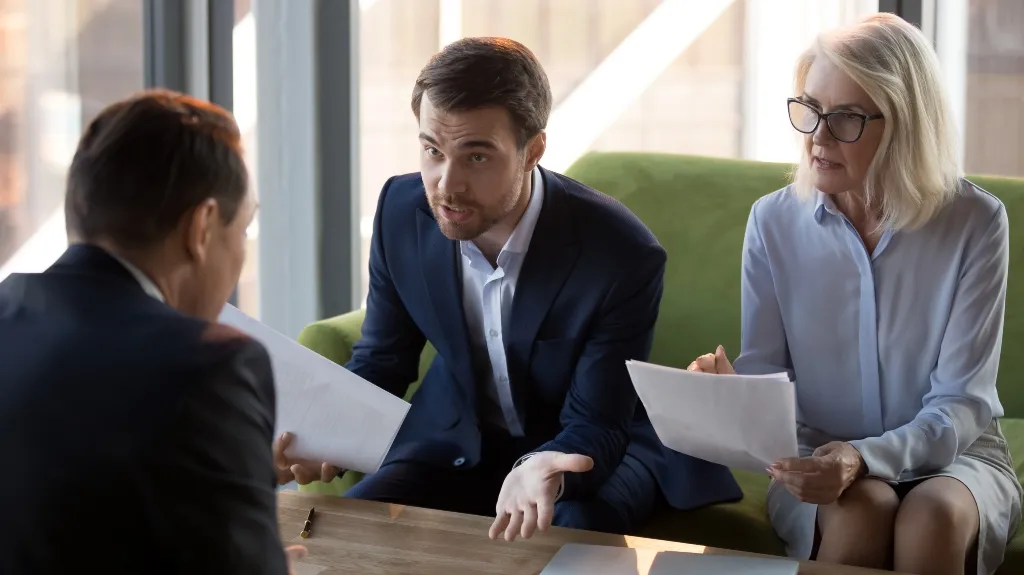 Three business professionals in formal attire engaged in a discussion, holding documents.