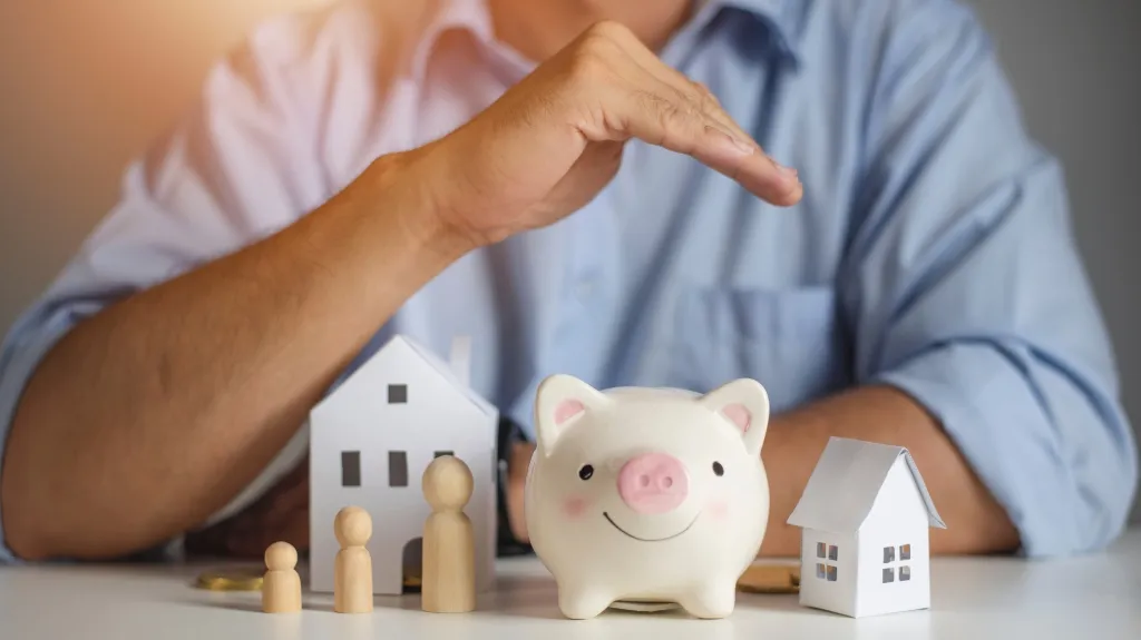 Person in blue shirt with hand protecting a white piggy bank, wooden family figures, and paper house models on table.